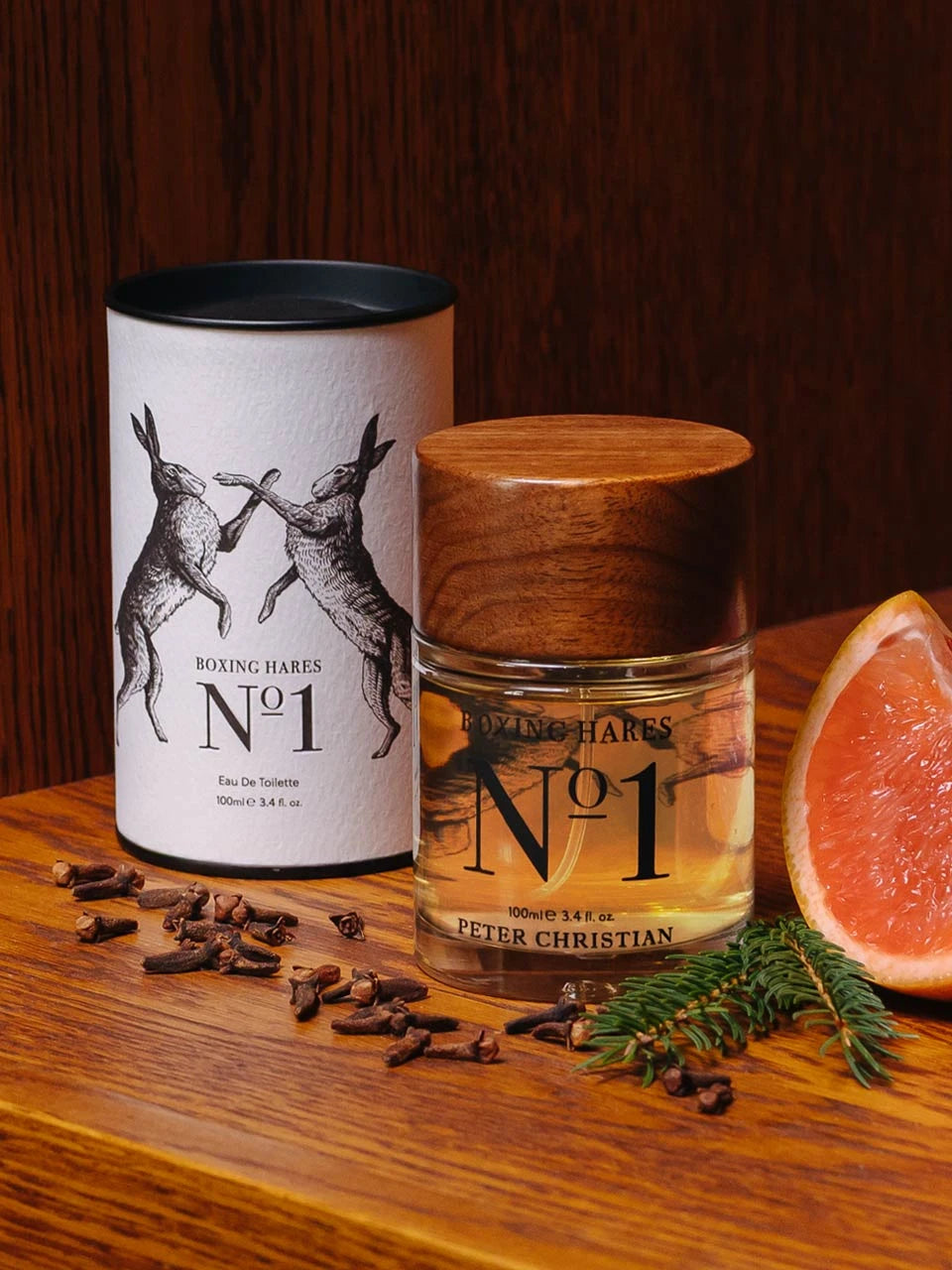 Peter Christian Boxing Hares No 1 Eau De Toilette shown with its white cylindrical tin and clear glass bottle with a wooden lid, styled on a wooden surface with cloves, pine sprigs, and a grapefruit slice.