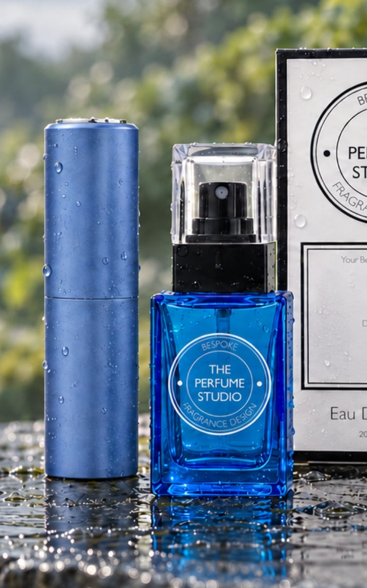 A blue rectangular perfume bottle labeled 'THE PERFUME STUDIO' stands in front of a matching blue cylindrical container and a branded box, all with water droplets, on a wet reflective surface with a blurred outdoor background.