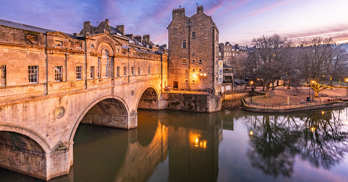 A scenic twilight photograph of the historic city of Bath an old stone bridge over calm water, with warm lights reflecting on the surface and bare trees along the riverbank under a pink and blue evening sky.