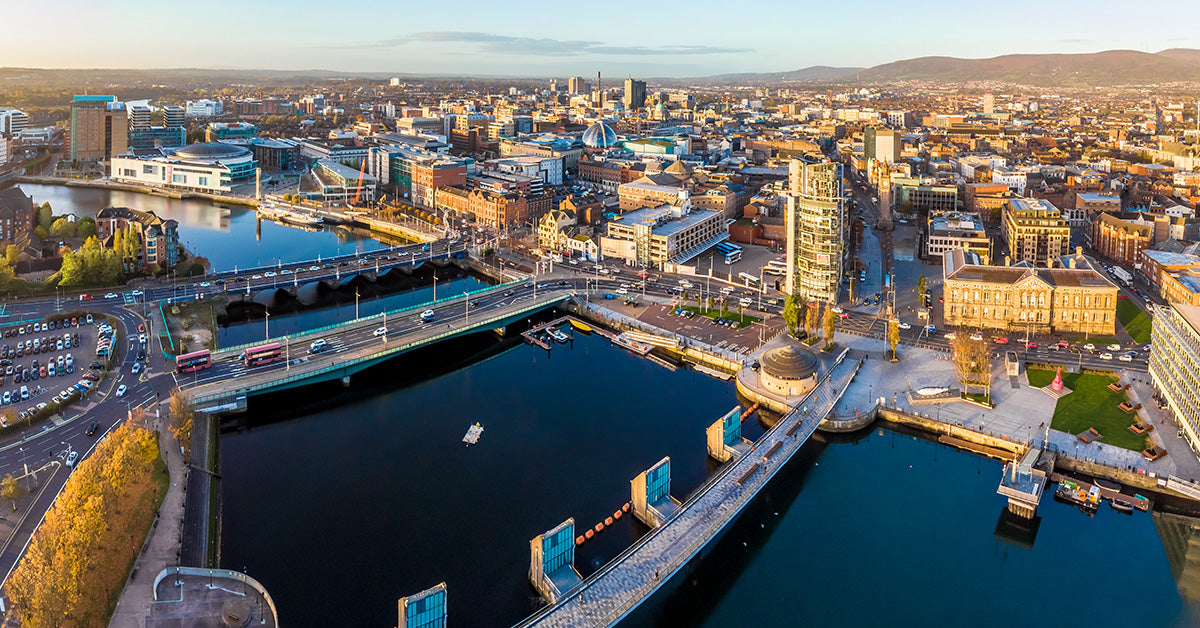 Aerial view of Belfast city waterfront with bridges over dark water, modern and historic buildings, roads, and distant hills under a clear sky.