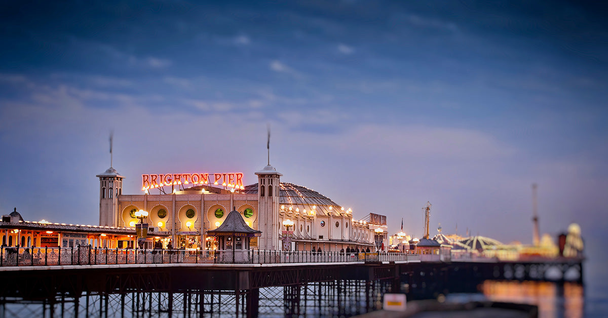 Brighton Pier at dusk with illuminated buildings, a large red 'BRIGHTON PIER' sign, and reflections on the water under a blue evening sky.