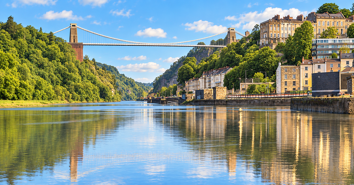 A scenic landscape of Bristol includes Clifton Suspension Bridge spanning Avon Gorge and River Avon, with lush green hills on one side and a row of historic buildings on the other, all reflected in the calm water under a blue sky with white clouds