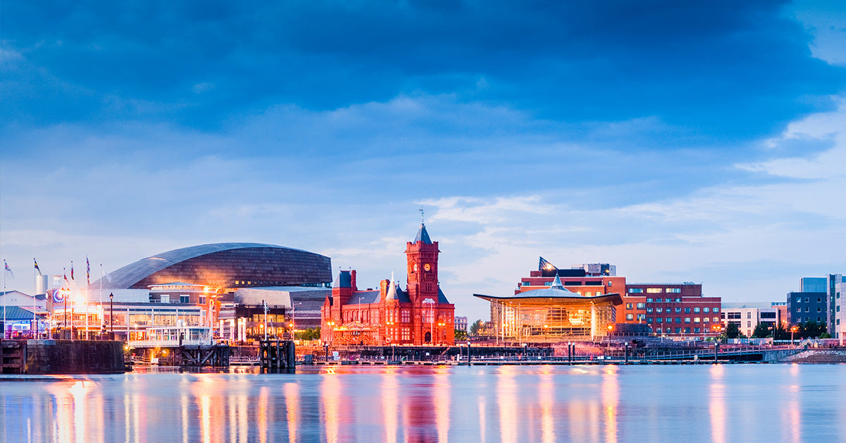 Cardiff waterfront city skyline at dusk with illuminated red-brick buildings, a modern dome structure, and reflections in calm water under a deep blue sky.