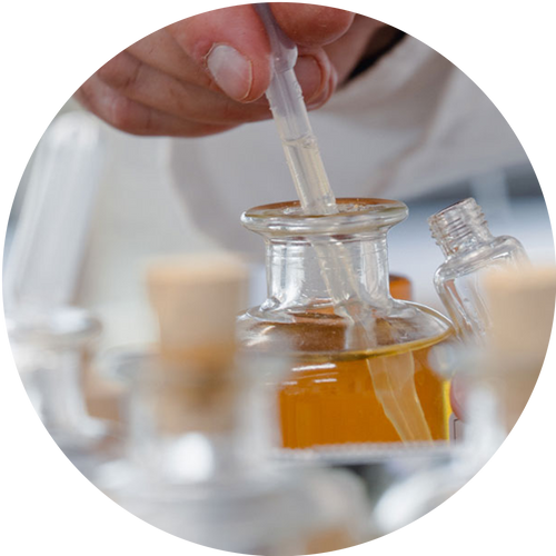 A close-up circular shot of a hand using a clear plastic pipette to transfer liquid into a small glass bottle containing amber liquid, with other glass bottles blurred in the background.