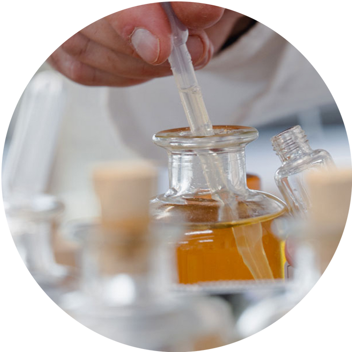 A close-up circular shot of a hand using a clear plastic pipette to transfer liquid into a small glass bottle containing amber liquid, with other glass bottles blurred in the background.