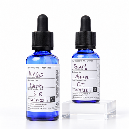 Two small cobalt blue glass dropper bottles with black caps stand on a white studio background, with one bottle placed on a clear acrylic riser. Both bottles have white handwritten-style labels with text including 'Your bespoke fragrance' and scent names 'Virgo' and 'Snap!'.