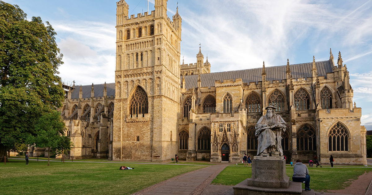 A wide outdoor view of a large historic Exeter cathedral with tall towers and pointed Gothic windows, set among green lawns and trees under a blue sky with light clouds. A statue of Richard Hooker stands in the foreground near a paved walkway.