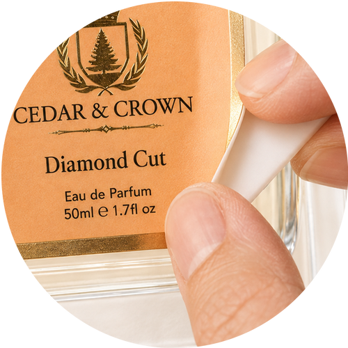 A close-up of a hand holding a white plastic dropper over a clear glass bottle labeled Cedar & Crown Diamond Cut Eau de Parfum, with a peach-colored label and gold text.