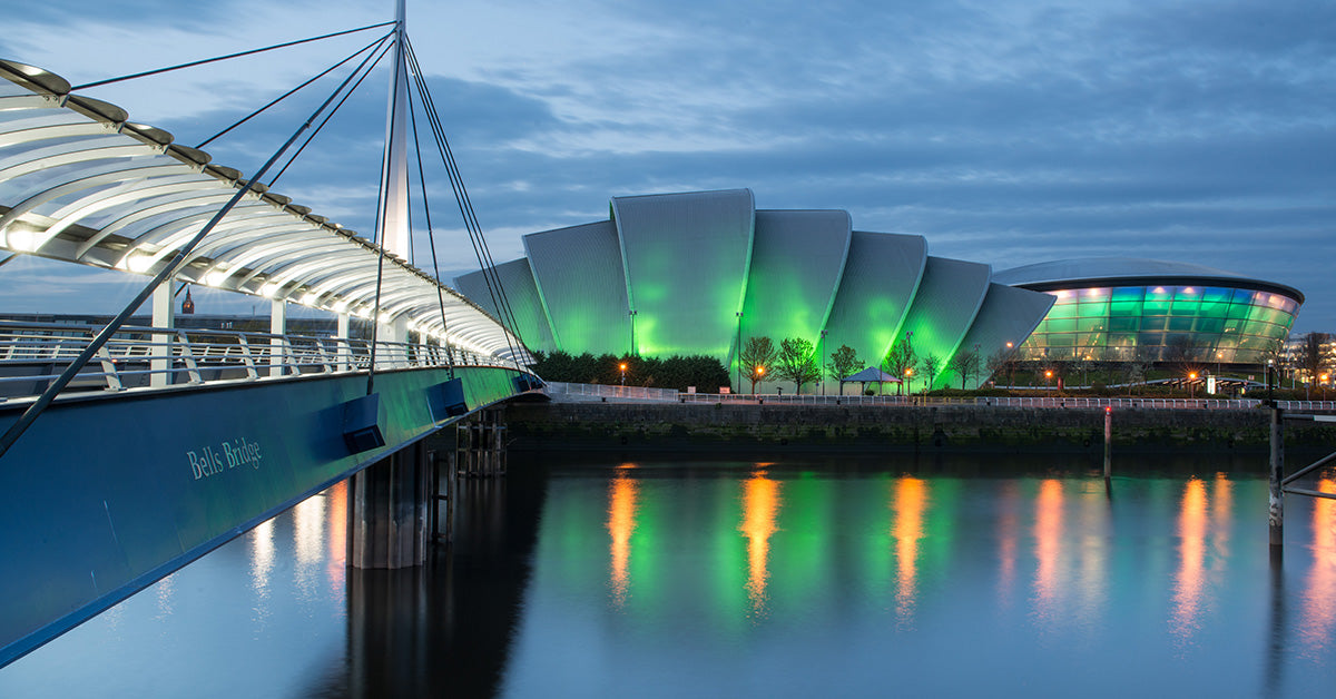 A twilight cityscape over Bells Bridge with a modern illuminated stadium and bridge reflected in calm water. The architecture is lit in green and blue tones against a deep blue evening sky.