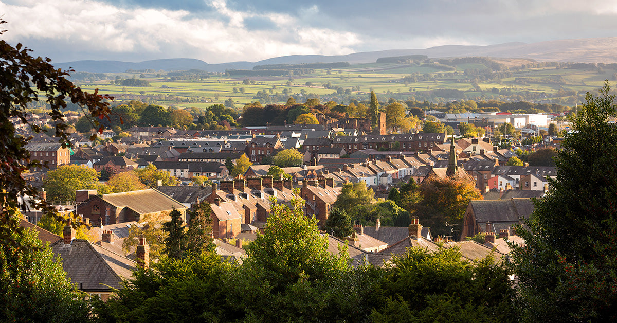 A scenic panoramic view of Penrith town in The Lake District with dense rooftops, trees, and rolling hills in the background under a cloudy sky.