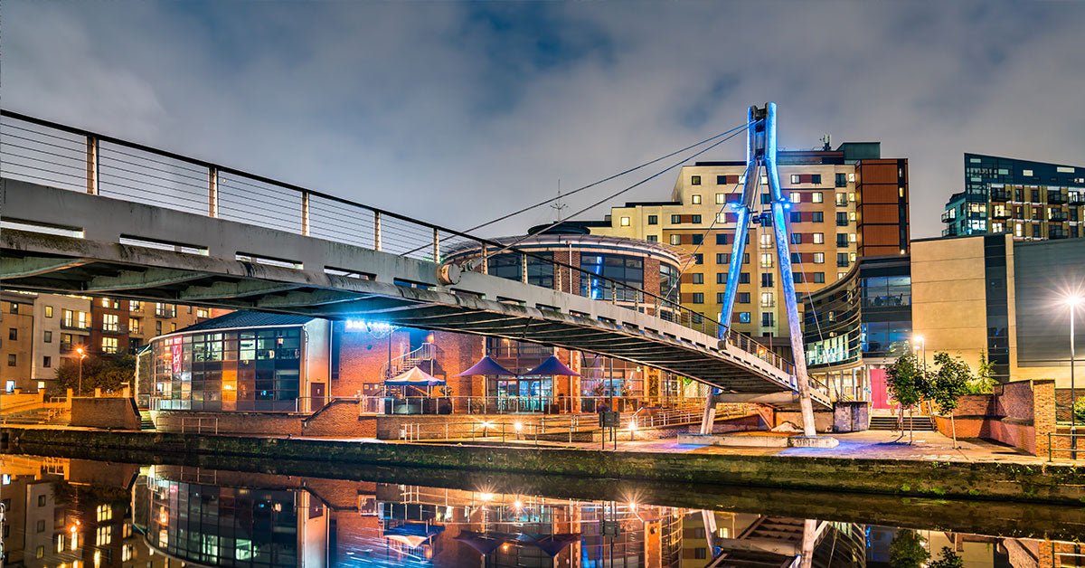Centenary Bridge, Leeds. A modern suspension footbridge spans a canal at night, with illuminated buildings and blue accent lighting on the bridge structure. The scene is reflected in the water below, with urban architecture visible in the background.
