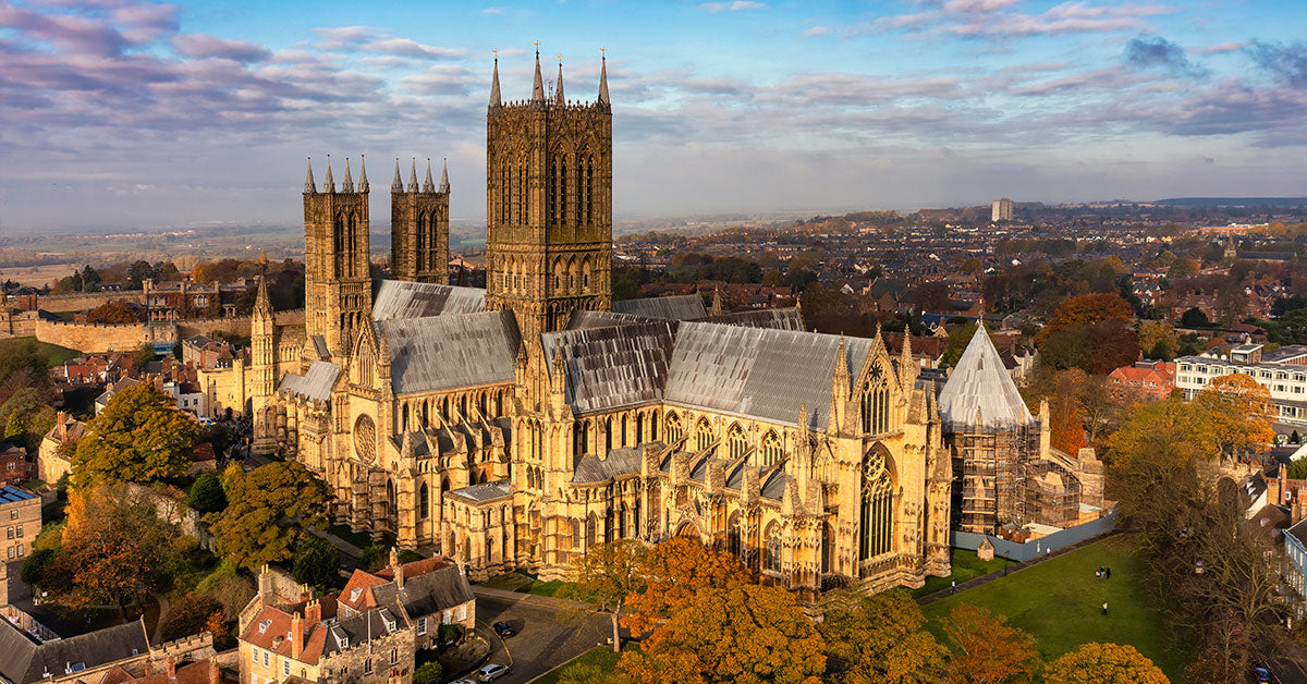Aerial view of a large historic cathedral in Lincoln with tall stone towers, surrounded by autumn trees and a town under a blue sky with scattered clouds.