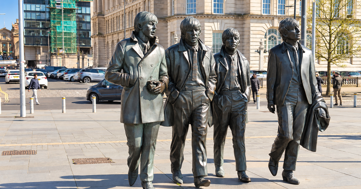 A bronze statue of the Beatles walking together in Liverpool, an outdoor urban plaza, with a large classical building and parked cars in the background.