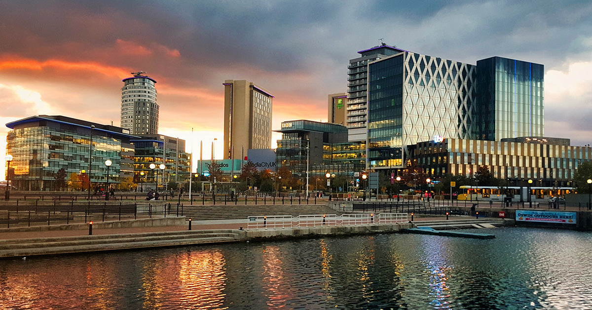 Manchester city skyline at sunset with modern glass buildings reflected in calm water, under a dramatic cloudy sky with orange and blue tones.