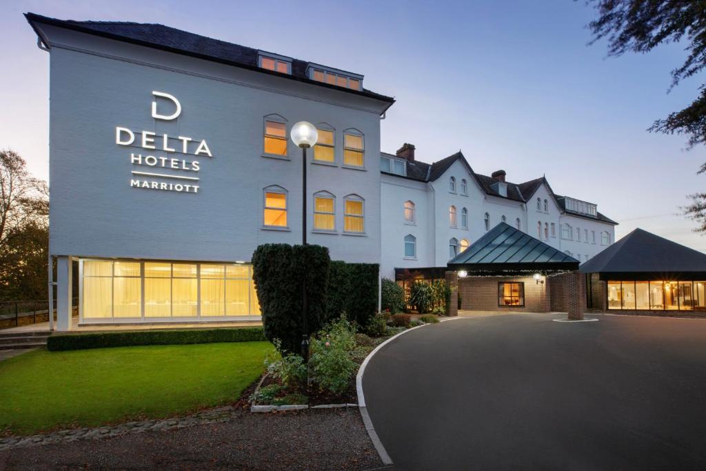 Delta Hotels Marriott hotel , York, exterior at dusk, showing a large light gray building with illuminated windows, a curved driveway, landscaped lawn, and entrance canopy.