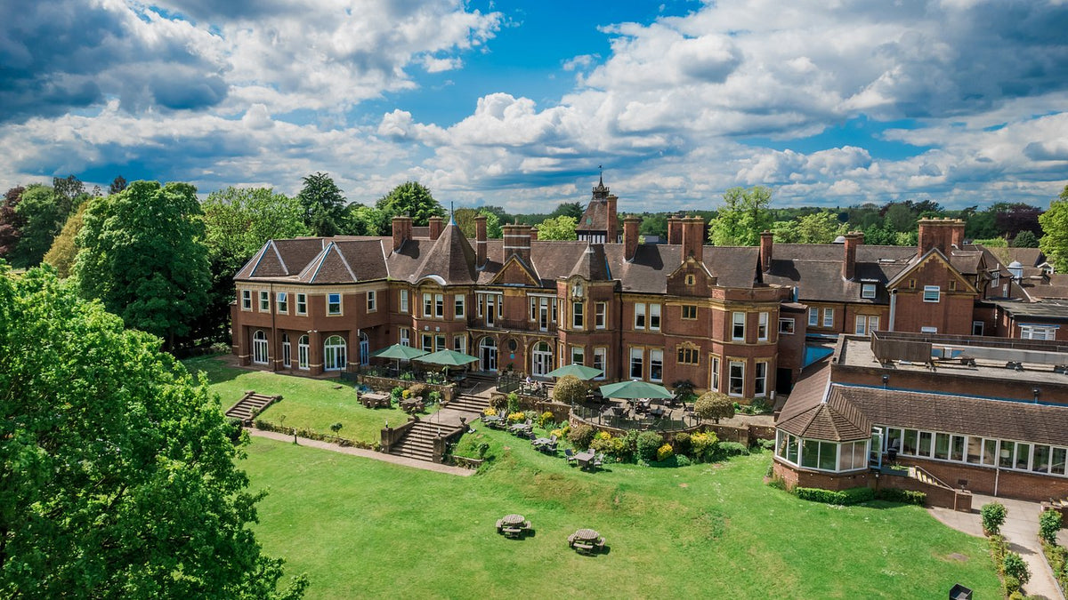 Aerial view of Moor Hall Hotel and Spa, Sutton Coldfield, Birmingham. A large red brick estate mansion with extensive green lawns, trees, outdoor patio umbrellas, and a dramatic blue sky with white clouds.