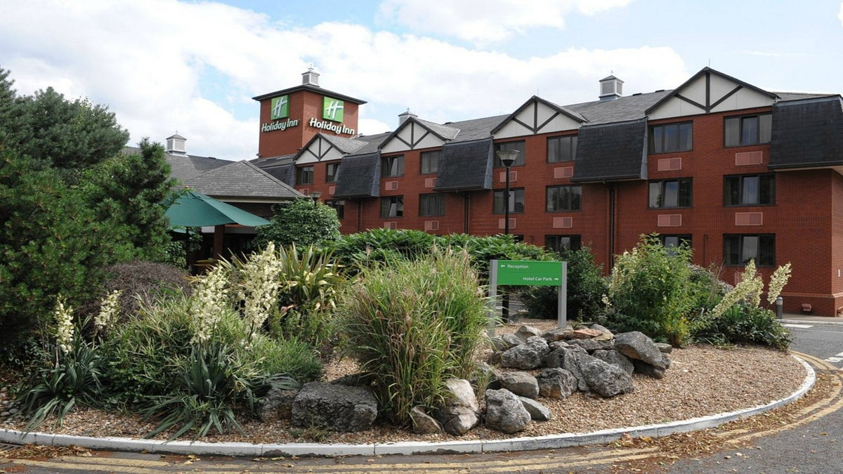 Holiday Inn Express, Northampton, hotel building with red brick exterior, dark gray roof, landscaped garden with rocks and shrubs, and a cloudy sky background.