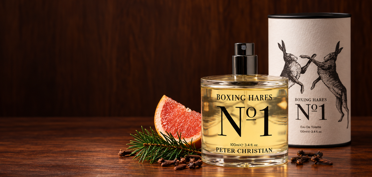 Peter Christian Boxing Hares No 1 Eau de Toilette perfume bottle and its cylindrical cardboard packaging, styled on a dark wooden table with a grapefruit slice, pine needles, and cloves.