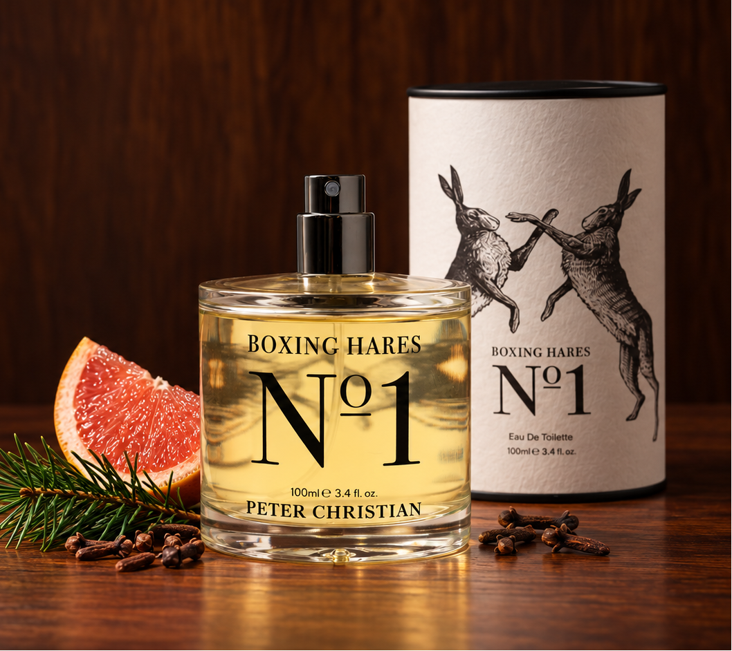 Peter Christian Boxing Hares No 1 Eau de Toilette perfume bottle and its cylindrical cardboard packaging, styled on a dark wooden table with a grapefruit slice, pine needles, and cloves.