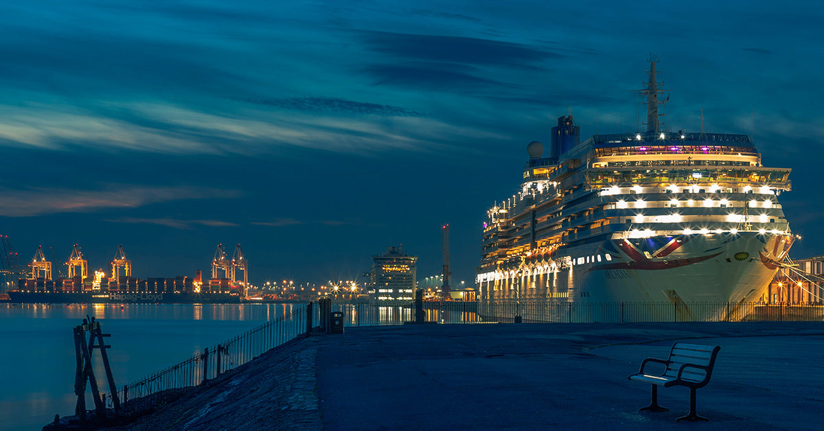A large illuminated cruise ship docked at Southampton harbor at dusk, with a pier in the foreground, a bench, and industrial buildings lit up in the distance under a deep blue evening sky.
