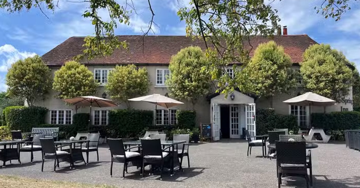 The Spring Inn at Sulhastead, a large stone building with a red-tiled roof, white-framed windows, and a gravel patio in front with outdoor dining tables and chairs under beige umbrellas, surrounded by green trees under a blue sky.