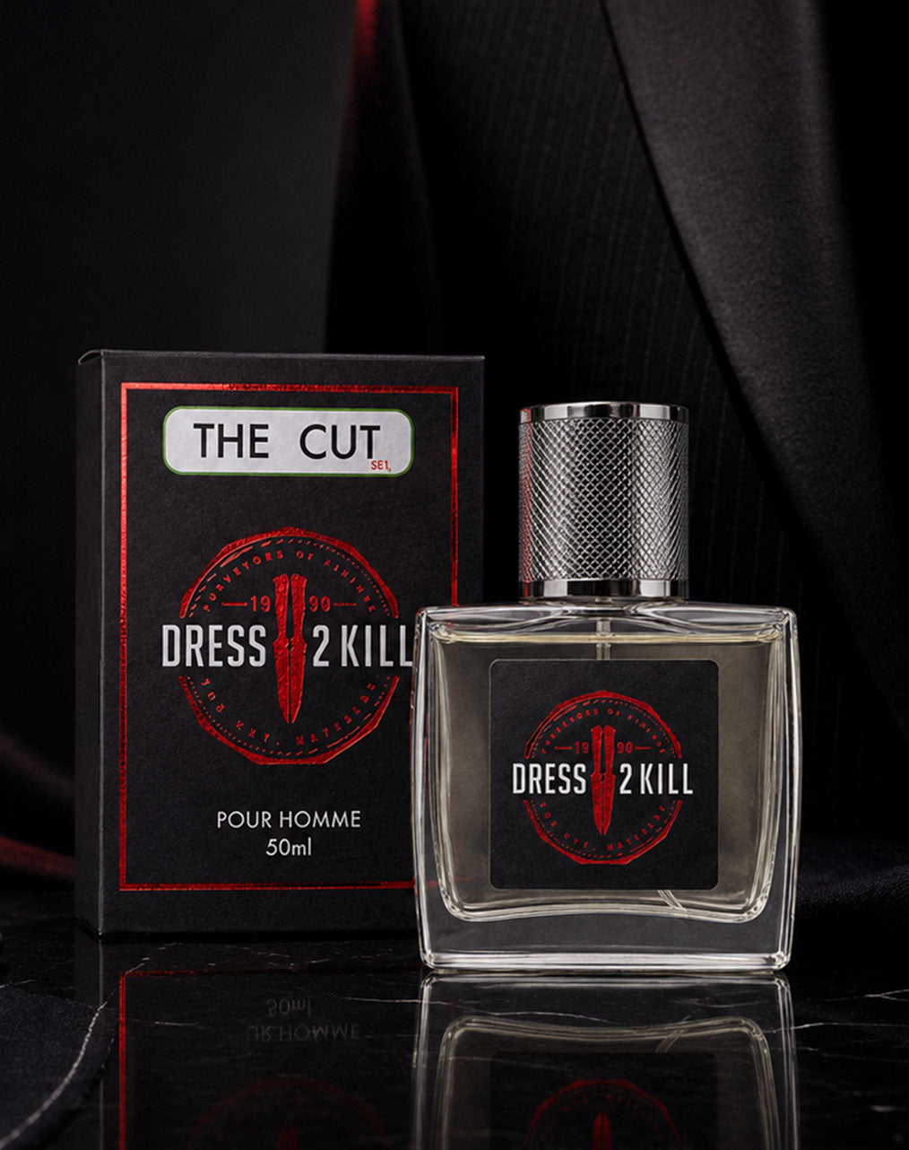 A bottle of Dress 2 Kill 'The Cut' perfume for men next to its black packaging box, styled on a dark reflective surface with a black pinstripe jacket in the background.
