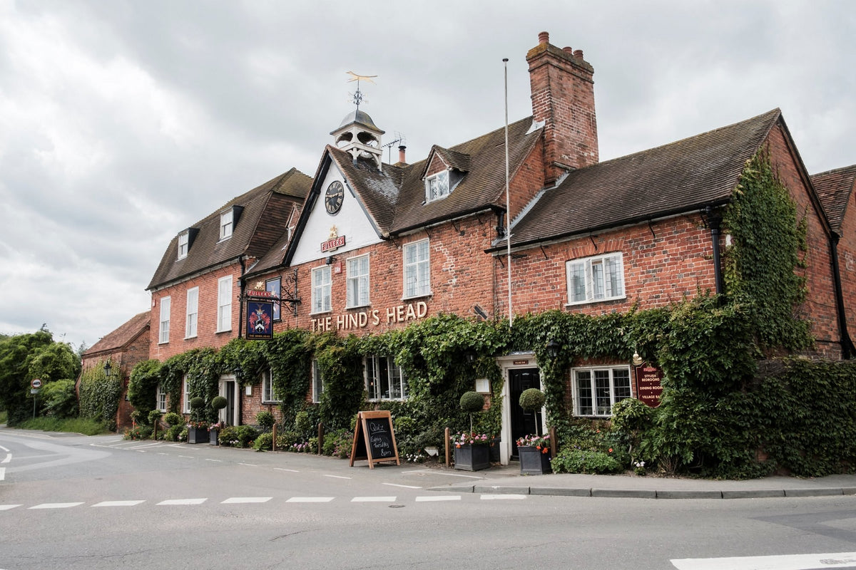 The Hind's Head pub exterior in Aldermaston, Reading. A village setting, featuring a red brick building with white-framed windows, a green ivy-covered facade, and a cloudy sky overhead.
