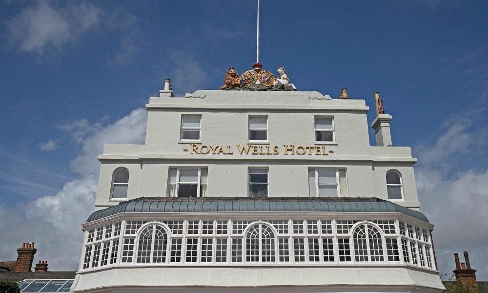 The front facade of the Royal Wells Hotel in Tunbridge Wells, featuring a white building with gold lettering, a glass conservatory on the ground floor, and a blue sky with clouds in the background.