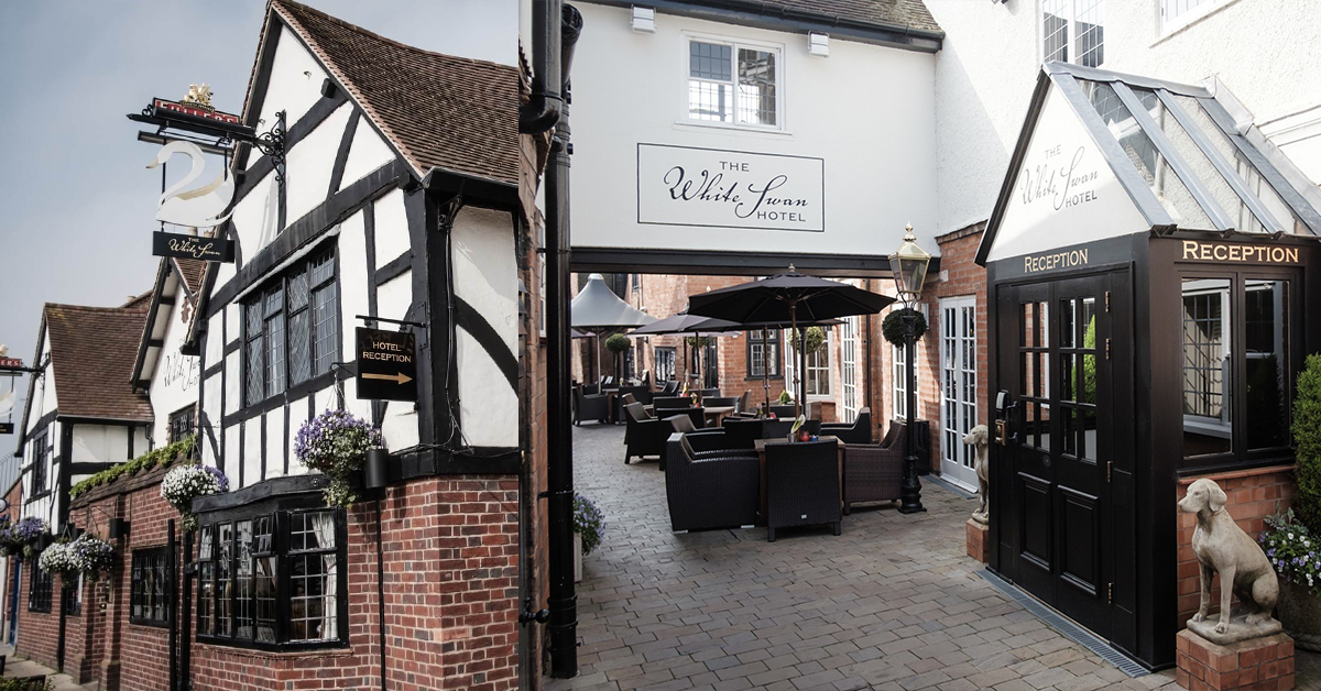 A wide exterior view of a historic hotel in Stratford-upon-Avon with timber-framed buildings, a brick chimney, outdoor seating under umbrellas, a white sign reading 'The White Lion', and a black reception booth with a dog statue beside it.